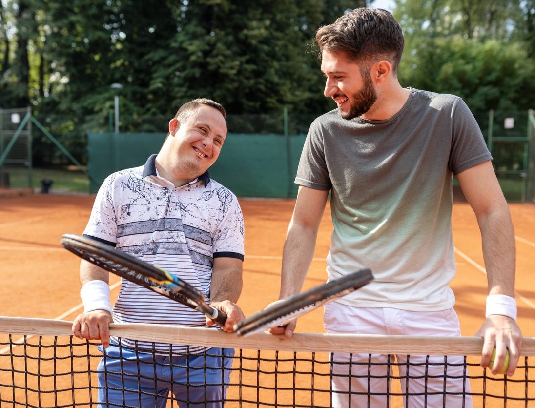 Young man with Down syndrome playing tennis with his friend on clay tennis court